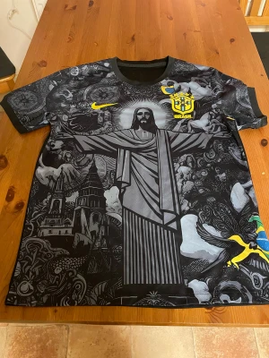 Brasil svart fotbollströja Nike - Unik svart Brasilien fotbollströja från Nike med detaljerat grafiskt tryck av Kristusstatyn och brasilianska motiv på framsidan. Gula Brasil-loggan och Nike swoosh på bröstet. Kortärmad modell i lätt, ventilerande material. Perfekt för dig som vill sticka ut på planen.