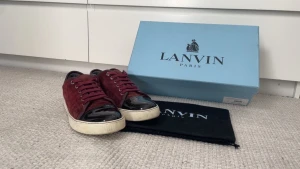 Lanvin bytes  - Säljer ett par snygga vinröda sneakers från Lanvin Paris med ovandel i mocka och tåparti i svart lack. Skorna har rund tå, platt vit sula och matchande vinröda snören. Kommer med originalkartong och dustbag. Perfekt för dig som gillar exklusiva detaljer.