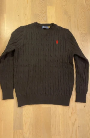 Svart kabelstickad tröja Polo Ralph Lauren - Snygg svart stickad tröja från Polo Ralph Lauren med klassiskt kabelstickat mönster och röd broderad logga på bröstet. Rund hals och långa ärmar, perfekt för dig som gillar stilrena och tidlösa plagg. Denna är äkta köpt på Zalando för 2200