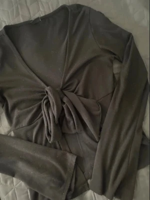 Svart omlottopp med knyt framtill - Säljer en svart omlottopp med långa ärmar och snygg knytning framtill som ger en cool croppad look. Toppen har v-ringad hals och är gjord i mjukt material som sitter skönt mot kroppen. Perfekt att styla med jeans eller kjol för en trendig vibe.