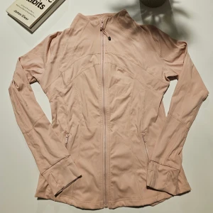 Ljusrosa vindjacka från Lululemon - Snygg ljusrosa vindjacka från Lululemon med hel dragkedja framtill och diskret logga på ryggen. Jackan har långa ärmar, figurnära passform och praktiska fickor med dragkedja. Perfekt för träning eller chill dagar ute.