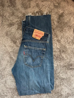 Levi's 501 blå jeans med rå kant - Klassiska Levi's 501 jeans i blå tvätt med rak passform och hög midja. Jeansen har fem fickor, ikonisk röd Levi's-tag på bakfickan och rå, fransad kant nedtill för en cool vibe. Tillverkade i slitstarkt bomullsjeansmaterial.Supersnygga och sköna! Midjemått: 40cm. Innerbenslängd: 72cm