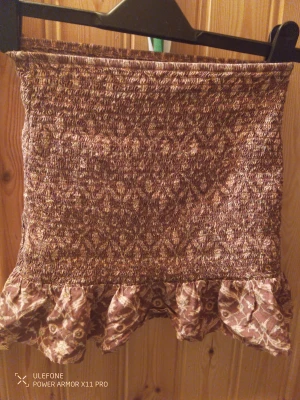 Brun mönstrad kortkjol från VNG - Säljer en brun kortkjol från VNG med ett unikt mönster och volangkant längst ner. Kjolen har en smockad midja som ger en skön och flexibel passform. 