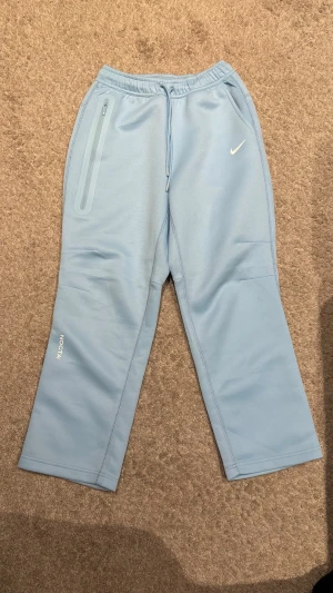 Nike NOCTA ljusblå mjukisbyxor - Nike NOCTA mjukisbyxor i ljusblått med snyggt tryck på benet och klassisk Swoosh-logga. Byxorna har snörning i midjan, en dragkedjeficka och är tillverkade i mjukt syntetmaterial. Perfekt för chill eller träning, loose fit och stilren design.