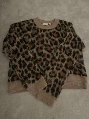Leopardmönstrad stickad tröja Kappahl XS - Säljer en fluffig stickad tröja från Kappahl i storlek XS. Tröjan har ett snyggt leopardmönster i beige, brun och svart med ribbade muddar vid hals, ärmslut och nederkant. Perfekt för dig som gillar djurmönster och vill ha en mysig look.