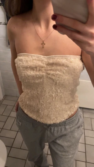 Champagnefärgad paljett tubtopp - Säljer en champagnefärgad tubtopp med glittriga paljetter över hela ytan. Toppen är axelbandslös och har en bandeauform som sitter tight mot kroppen. Perfekt för dig som vill sticka ut med lite extra bling. Materialet känns syntetiskt och skimrar snyggt i ljuset. Passar s-m. Perfekt för nyår!