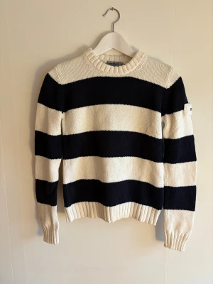 Randig stickad tröja Henri Lloyd - Snygg stickad tröja från Henri Lloyd med breda marinblå och vita ränder. Tröjan har rund hals, ribbade muddar och ett broderat märke på ärmen. Perfekt för dig som gillar klassisk och sportig stil. Materialet är mjuk bomull. Tröjan har krymt lite i tvätten.