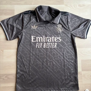 Real Madrid grå matchtröja Adidas - Säljer en grå Real Madrid t-shirt från Adidas med korta ärmar och krage. Tröjan har ett diskret mönster, klubbmärke och Emirates-tryck på bröstet. Materialet är polyester och modellen är normal passform. Perfekt för fotbollsfans!