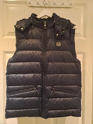 Mörkblå dunväst från Bvaldi, storlek L - Mörkblå dunväst från Bvaldi med huva och två fickor med dragkedja. Nypris: 1799kr. Mycket fint skick.