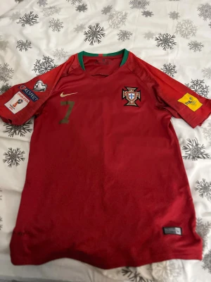 Portugal Ronaldo 7 Nike fotbollströja - Säljer en officiell Portugal fotbollströja från Nike med Ronaldo och nummer 7 på ryggen. Tröjan är röd med gröna detaljer vid kragen, har korta ärmar och tryckta märken på båda ärmarna. Materialet är lätt och andas, perfekt för match eller träning.