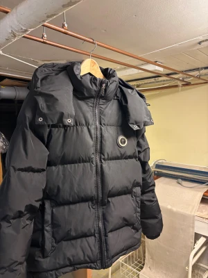 Trapstar svart pufferjacka med huva - Svart pufferjacka från Trapstar med stor broderad logga på ryggen och rund patch på bröstet. Jackan har huva, dragkedja framtill och två sidofickor. Puffad design och långärmad, perfekt för kalla dagar. Snygg streetstil med detaljerade broderier.