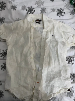 Vit linneskjorta från Morris - Fräsch vit skjorta från Morris i mjukt linne, med korta ärmar och klassisk krage. Skjortan har knappar framtill och en liten broderad logga på bröstet. Perfekt för varma dagar och en chill stil.