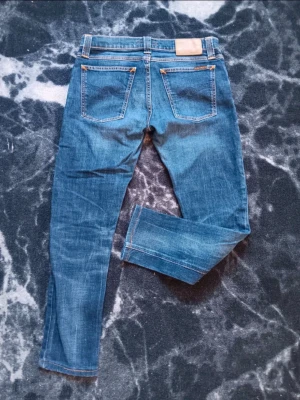 Blå jeans från Nudie Jeans Co - Snygga blå jeans från Nudie Jeans Co med klassisk femficksdesign och orange kontrastsömmar. Jeansen har rak passform och normal midja, tillverkade i slitstarkt denim med detaljerad logotyp på bakfickan och knapp. Perfekt för dig som gillar stilrena och tidlösa jeans.
