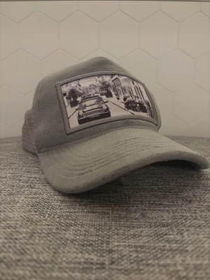 Grå keps Emilio Pendenza med bilmotiv - Snygg grå keps från Emilio Pendenza med mesh på baksidan och justerbar snapback. Framtill finns ett svartvitt motiv med en klassisk bil och stadsmiljö. Kepsen har en böjd skärm och är tillverkad i syntetmaterial med tygdetaljer.