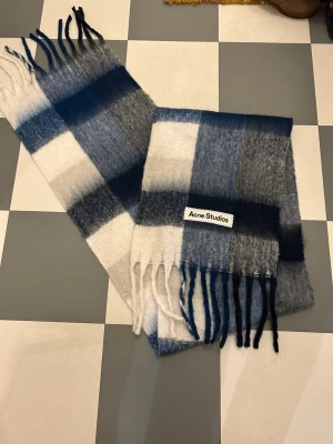 Rutig blå och vit halsduk Acne Studios - Snygg och fluffig halsduk från Acne Studios i rutigt mönster med blå, grå och vita toner. Mjuk och lyxig känsla, tillverkad i Italien. Långa fransar i ändarna och stor logotyp på ena sidan. Perfekt för att ge din outfit en cool touch. Nytt skick och priset är inte hugget i sten 