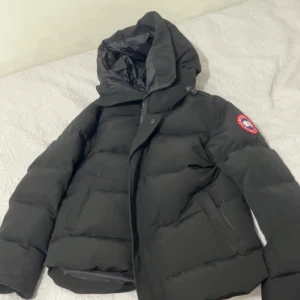 Svart dunjacka från Canada Goose - XS men passar S. Svart dunjacka från Canada Goose med huva och klassisk logga på ärmen. Jackan har en puffig design, dragkedja och knappar framtill. Perfekt för kalla dagar med sin tjocka och isolerande fyllning. Snygg och stilren look som funkar till det mesta.