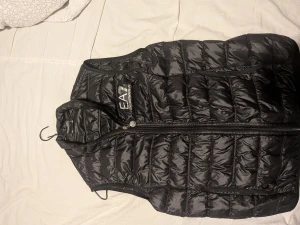Svart dunväst från EA7 Emporio Armani - Snygg svart dunväst från EA7 Emporio Armani med quiltad design och dragkedja framtill. Västen har en stilren look med logga på bröstet och är tillverkad i glansig polyester. Perfekt för lager-på-lager och streetstyle.