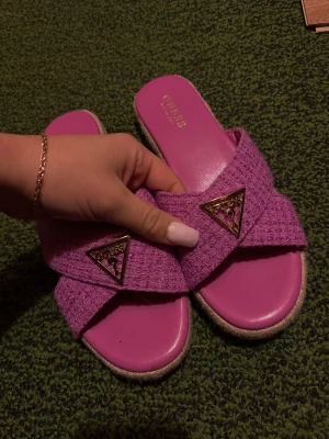 Rosa sandaler från Guess - Jag säljer mina snygga rosa sandaler från Guess med breda korslagda band i stickat material och guldiga logodetaljer framtill. Sandalerna har platt sula och öppen tå, perfekt för sommarens alla dagar. 