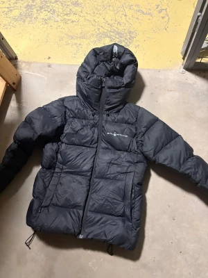 Svart pufferjacka från Sail Racing - Svart pufferjacka från Sail Racing med huva och dragkedja framtill. Jackan har ett quiltat mönster och logga på bröstet. Perfekt för kalla dagar med sin tjocka och fluffiga design. Justerbara snören nedtill och långärmad modell.