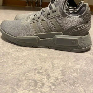 Grå Adidas NMD sneakers - Säljer ett par grå Adidas NMD sneakers med stickad ovandel och klassiska tre ränder på sidan. Skorna har en chunky sula med detaljer och snörning framtill. Perfekta för dig som gillar en sportig och modern look. Adidas-logga på sidan och plös. Helt nya och rena, inga fläckar alls. Haft på mig de några gånger. Nypris runt 1,7 tusen. Storlek 40 1/2 men jag har storlek 42 och de passar perfekt.