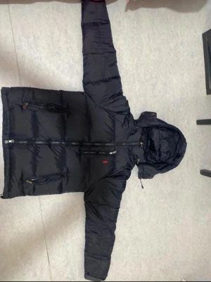 Svart dunjacka från Polo Ralph Lauren - Svart dunjacka från Polo Ralph Lauren med huva, dragkedja och tre fickor med zip framtill. Jackan har det klassiska röda Polo-logot broderat på bröstet och quiltad design. Perfekt för kalla dagar och har en snygg, normal passform.om du har några frågor som t:ex om priset eller något annat så kan du absoulut fråga.