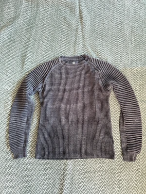 Grå stickad tröja G-Star RAW  - Snygg grå stickad tröja från G-Star RAW i storlek S. Tröjan har ett grovt stickat mönster på kroppen och ribbade, randiga ärmar som ger en cool kontrast. Rund hals och långärmad modell, perfekt för dig som gillar streetwear med lite edge.