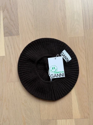 Mörkbrun stickad basker från Ganni - Snygg mörkbrun stickad basker från Ganni, tillverkad i Italien. Baskern har ett ribbstickat mönster och är gjord i mjukt ullmaterial. Perfekt för dig som vill ha en cool och trendig accessoar med lite Paris-vibe.