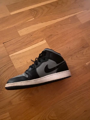 Nike Air Jordan 1 Mid Svart/Grå - Säljer ett par Nike Air Jordan 1 Mid i svart och grått skinn med klassisk svart swoosh och röda detaljer på hälen och plösen. Skorna har snörning, vit mellansula och svart yttersula. Perfekta för dig som gillar streetstyle och ikoniska sneakers. pris kan diskuteras😊
