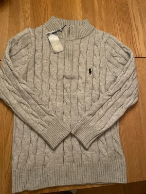 Grå stickad half zip tröja Polo Ralph Lauren - Snygg grå kabelstickad tröja från Polo Ralph Lauren med half zip och ribbad krage. Klassisk broderad logga på bröstet. Långärmad och tillverkad i mjukt stickat material som är perfekt för kyliga dagar. Passar dig som gillar stilren och tidlös look. Helt ny, Nypris 1500-2000, priset kan diskuteras vid snabb affär! Mvh Gustav 