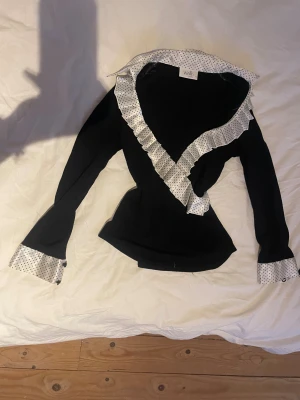 Svart tröja med prickiga detaljer  - Svart långärmad tröja från Wallis med vit krage, manschetter och volangkant i satin med svarta prickar. Snygg v-ringning och dubbla knappar på ärmsluten. Figurnära passform och cool kontrast mellan material och mönster.