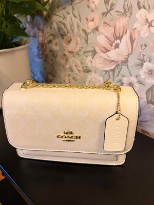 Vit axelväska från Coach med kedja - Snygg vit axelväska från Coach med guldfärgad kedja och logga framtill. Nypris 3000+ säljer för 800kr Väskan har flera fack, svarta kanter och subtilt mönster med Coachs signatur. Materialet är skinn och den har en tag med Coach New York. Perfekt storlek för det viktigaste.