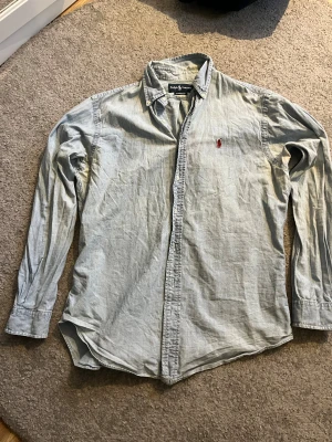 Ljusblå skjorta från Ralph Lauren M - Säljer en ljusblå klassisk skjorta från Ralph Lauren i storlek M. Skjortan har lång ärm, knappar framtill och den ikoniska röda broderade loggan på bröstet. Materialet är mjuk bomull och passformen är normal classic fit. Perfekt för dig som gillar stilrena plagg med snygg detalj.