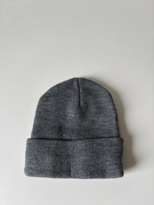 Grå stickad mössa  - En enkel och stilren grå stickad mössa med uppvikt kant. Perfekt för kalla dagar och passar till de flesta outfits. Mjuk och skön, håller dig varm och ser snygg ut samtidigt. Helt ny.