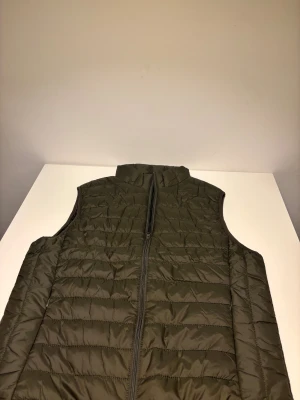 Mörkgrön quiltad dunväst - Snygg mörkgrön dunväst med quiltad design och hög krage. Västen har en rak passform och stängs med dragkedja framtill. Perfekt att slänga över en hoodie för extra värme. Materialet är lätt och smidigt, vilket gör den enkel att bära och styla. Helt ny.