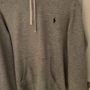 Grå hoodie från Polo Ralph Lauren - Snygg grå hoodie från Polo Ralph Lauren med klassisk logga på bröstet. Tröjan har huva med vita snören, stor magficka och mjukt material som känns skönt mot huden. Perfekt för chill dagar och enkel att matcha med jeans eller joggers.