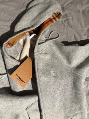 Grå hoodie från Burberry med dragkedja - Säljer en grå hoodie från Burberry med klassiskt rutigt foder i huvan och broderad logga på bröstet. Tröjan har dragkedja framtill, snörning i huvan och är fodrad för extra komfort. Perfekt för dig som vill ha en stilren och lyxig look.