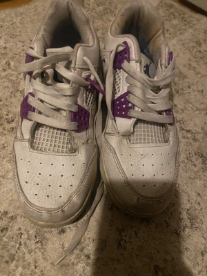 Nike Air Jordan 4 Retro vit/lila - Nike Air Jordan 4 Retro sneakers i vitt skinn med lila detaljer och meshpaneler. Klassisk Jumpman-logga på hälen och coola lila accenter vid snörningen. Platt sula och rund tå, perfekt för dig som gillar streetstyle och ikoniska sneakers.