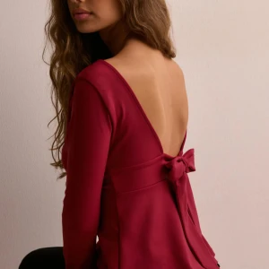 Röd långärmad topp med rosett - Helt ny!! Snygg röd topp med djup v-ringning och långa ärmar. Toppen har en elegant öppen rygg med en stor rosett som detalj. Perfekt för en stilfull look.