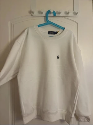 Vit sweatshirt från Polo Ralph Lauren - Vit sweatshirt från Polo Ralph Lauren med klassisk rund hals och broderad logga i svart på bröstet. Tröjan har långärmad passform och ribbade muddar vid ärmslut och nederkant. Perfekt för en clean och stilren look.