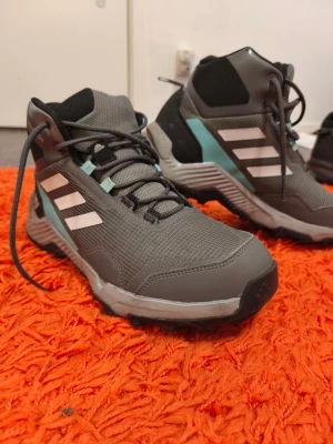 Grå höga sneakers från Adidas strl 40 - Säljer ett par grå höga sneakers från Adidas med vita ränder och svarta detaljer. Skorna har snörning, robust sula och inslag av ljusblått. Tillverkade i syntetmaterial och textil, perfekta för dig som gillar sportig stil.
