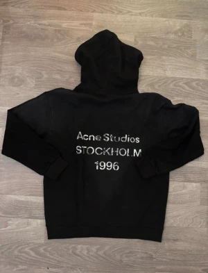 Svart hoodie Acne Studios Stockholm - Svart hoodie från Acne Studios med stor huva och tryck i silver på ryggen: 'Acne Studios STOCKHOLM 1996'. Tröjan har långa ärmar och ribbade muddar. Materialet är mjuk bomull, perfekt för chill dagar. Snygg och enkel design med tag kvar.