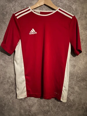 Röd Adidas fotbollströja Climalite - Snygg röd fotbollströja från Adidas med vita detaljer och klassiska tre ränder på axlarna. Tröjan har rund hals, korta ärmar och är tillverkad i lätt, ventilerande Climalite-material som håller dig torr under matchen. Adidas-logga på bröstet och vita paneler längs sidorna.
