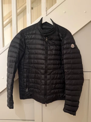 Svart Moncler dunjacka herr - Svart dunjacka från Moncler med quiltad design och klassisk logga på ärmen. Jackan har hög krage, dragkedja framtill och två sidofickor med dragkedja. Perfekt för kalla dagar och har en stilren, sportig look.