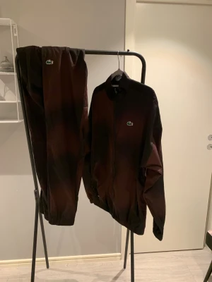 Lacoste tränings dress, storlek S  - Säljer ett par svarta mjukisbyxor från Lacoste med elastiska muddar och klassisk grön krokodillogga på benet. Materialet är mjukt och bekvämt, perfekt för chill dagar. Byxorna har en mörk ton och matchar snyggt med jackan i samma set.