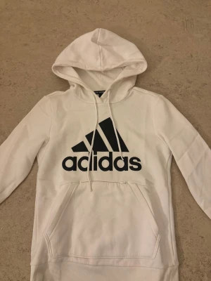 Vit hoodie från Adidas med logga - Säljer en vit hoodie från Adidas med stor svart logga på bröstet. Tröjan har huva med snörning, lång ärm och en klassisk magficka. Mjuk insida och ribbade muddar vid ärmslut och nederkant. Perfekt för chill eller sportiga dagar.