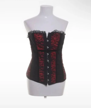 Svart och röd korsett med spetsdetaljer - Säljer en snygg korsett i svart och rött med spets upptill och dekorativa krokar framtill. Korsetten har ett blommigt mönster i rött och svarta paneler på sidorna. Axelbandslös design som ger en cool och edgy look.
