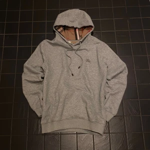 Grå burberry hoodie - Säljer en grå hoodie från Burberry med klassiskt rutigt foder i huvan och broderad logga på bröstet. Tröjan har snörning i huvan, ribbade muddar och en skön passform. Perfekt för dig som gillar stilrena och exklusiva plagg.