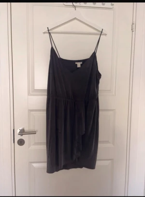 Svart omlottklänning från H&M - Svart kortklänning från H&M med tunna axelband och omlottdetalj framtill. Klänningen har en v-ringad hals och är tillverkad i ett mjukt, lätt tyg som känns skönt mot huden. Perfekt för dig som gillar en enkel men stilren look.