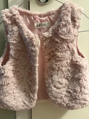 Ljusrosa pälsväst från H&M - Supermjuk och fluffig ljusrosa pälsväst från H&M. Västen är ärmlös och har en rundad halsringning med slät insida. Perfekt för att addera lite extra stil och värme till ditt barns outfit. Snygg till både klänning och långärmad tröja.