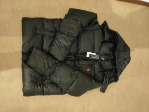 Svart dunjacka från Polo Ralph Lauren - Svart dunjacka från Polo Ralph Lauren med klassisk röd broderad logga på bröstet. Jackan har quiltad design, huva och dragkedja framtill. Perfekt för kalla dagar och har en clean, stilren look som funkar till allt.